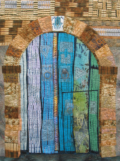 Tunisian Door, 106 cm x 140 cm, by Margaret Ramsay
Pieced from African fabrics including tie-dye and batik fabrics from Ghana and The Gambia with curved insert ‘gaps’ pieced from indigo and black waxprint . Other fabrics used were Kudinda Ndora Stripe, kola nut and indigo.  The ‘Hand of Fatima’ door knockers and rivet patterns were printed from photographs using ink jet printer onto cotton or silk organza treated with ‘Bubble Jet Set’. Door knockers were hand painted with inktense pencils to match the surrounding batik. Then the artist machine quilted the door with variegated threads.